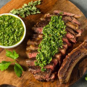 Sliced medium-rare steak topped with vibrant chimichurri sauce on a wooden board, served with a bowl of homemade chimichurri, fresh herbs, and a parsley garnish on the side.