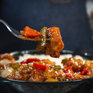 A close-up of a person holding a fork with a bite of saucy sweet and sour chicken and colorful vegetables above a bowl of the same dish, served with white rice. The perfect shot for any sweet and sour chicken recipe.