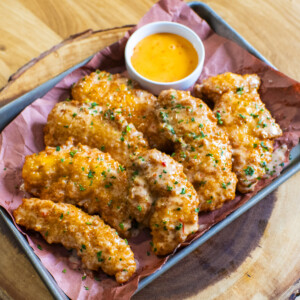 A tray of crispy fried chicken tenders, reminiscent of bang bang chicken, garnished with herbs on pink parchment paper and served with a small cup of creamy bang bang sauce on a round wooden board.