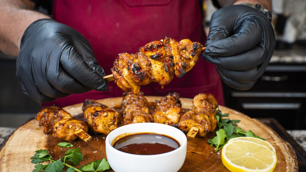 A person wearing black gloves holds a grilled chicken skewer over a bowl of dipping sauce, with more chicken skewers, parsley, and a lemon slice on a wooden board. The person wears a red apron.
