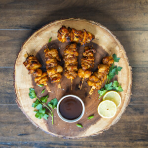 Grilled chicken skewers arranged on a round wooden platter with a small bowl of dipping sauce, fresh parsley, and lemon slices, set on a wooden table&mdash;perfect for those who love delicious chicken skewers cooked to perfection.