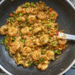 Shrimp fried rice with peas, carrots, and green onions being stirred in a skillet with a white spatula—this shrimp fried rice with cauliflower is perfect for a lighter twist, shown on a kitchen countertop next to a wooden cutting board and knife.