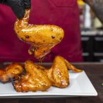 A person wearing black gloves and a red apron holds up a sweet and spicy chicken wing over a plate with more wings on a kitchen counter. Chopped green onions are visible on the side.