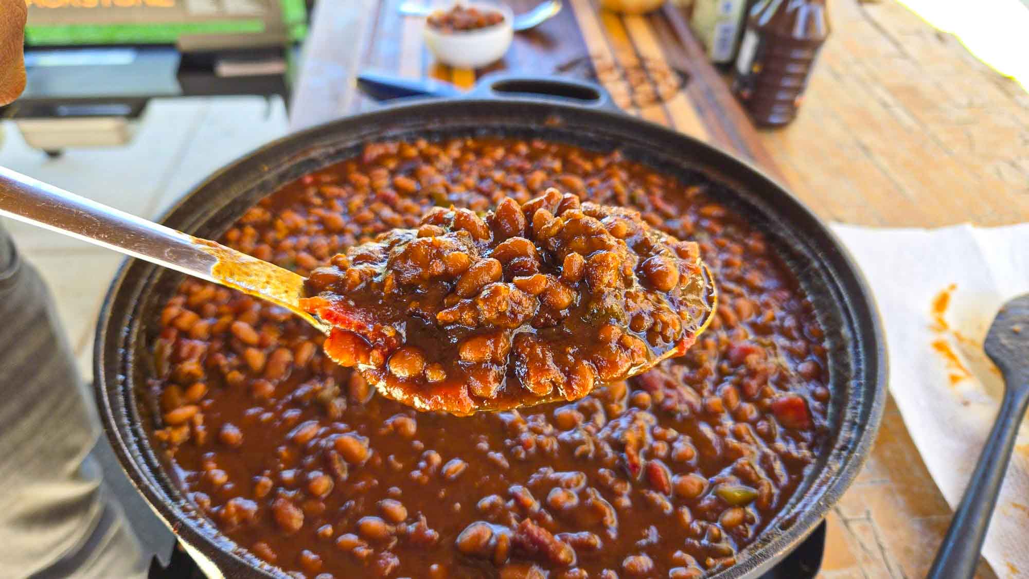 A close-up of a spoon holding BBQ baked beans in a thick sauce over a large pot, with more baked beans visible in the background on a wooden table.