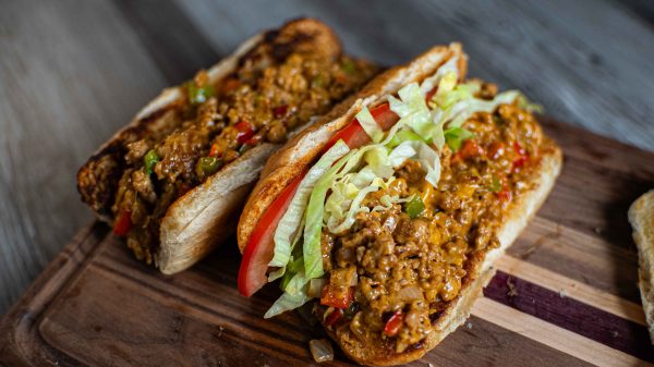 Two toasted sandwich rolls filled with seasoned ground meat, peppers, shredded lettuce, and tomato slices—a delicious twist on the classic chopped cheese sandwich—served on a wooden cutting board.