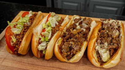Four hoagie buns filled with different toppings are lined up on a wooden board. Two have ground meat, lettuce, and tomato, while the others feature an authentic Philly cheesesteak recipe with chopped steak and melted cheese.