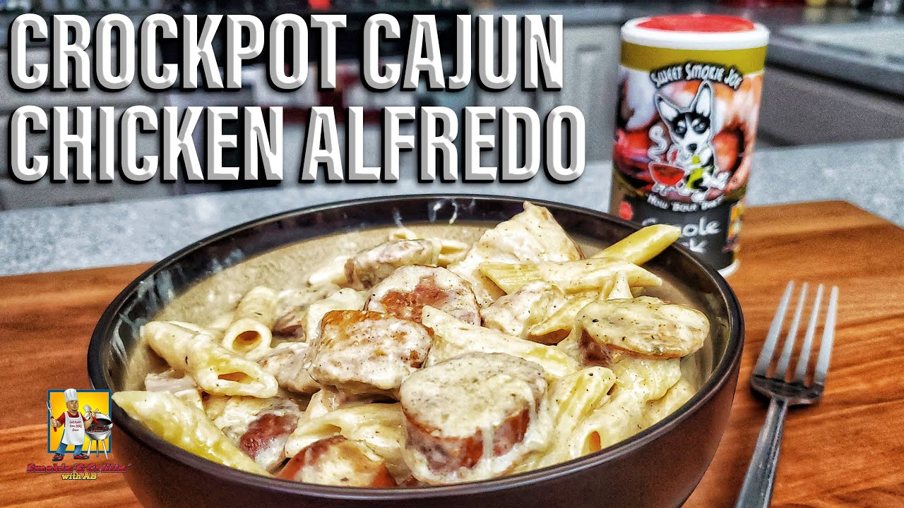 A bowl of creamy crockpot chicken alfredo with sliced sausage sits on a wooden surface. Behind it, a container of Cajun seasoning and large bold text reads Crockpot Cajun Chicken Alfredo. Try this easy crockpot chicken alfredo recipe!.