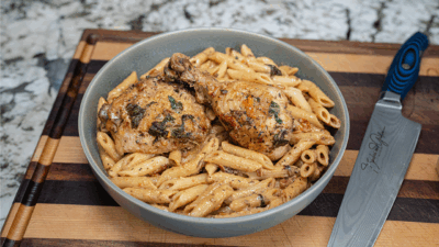 A bowl of creamy marry me chicken pasta, featuring penne topped with two seasoned drumsticks, sits on a striped wooden cutting board next to a chef's knife with a blue handle.
