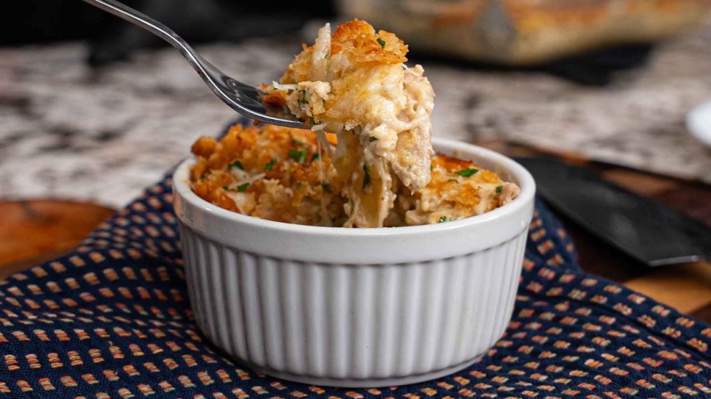 A fork lifts a cheesy, golden-brown serving of chicken casserole with Ritz crackers from a white ramekin, showcasing shredded chicken and a crispy topping. The ramekin sits on a patterned cloth on a kitchen counter.
