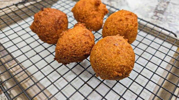 Five golden-brown hush puppies rest on a black wire cooling rack, set on a light-colored surface. These classic hush puppies appear crispy and round, perfectly showcasing their signature crunchy texture.