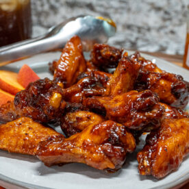 A plate of hot honey peach wings is served with apple slices. A pair of tongs, a jar of peach preserves, and a fresh peach are visible in the background on a patterned cloth.
