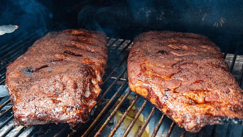 Juicy smoked ribs glazed with barbecue sauce, stacked on a wooden cutting board beside a bowl of sauce.