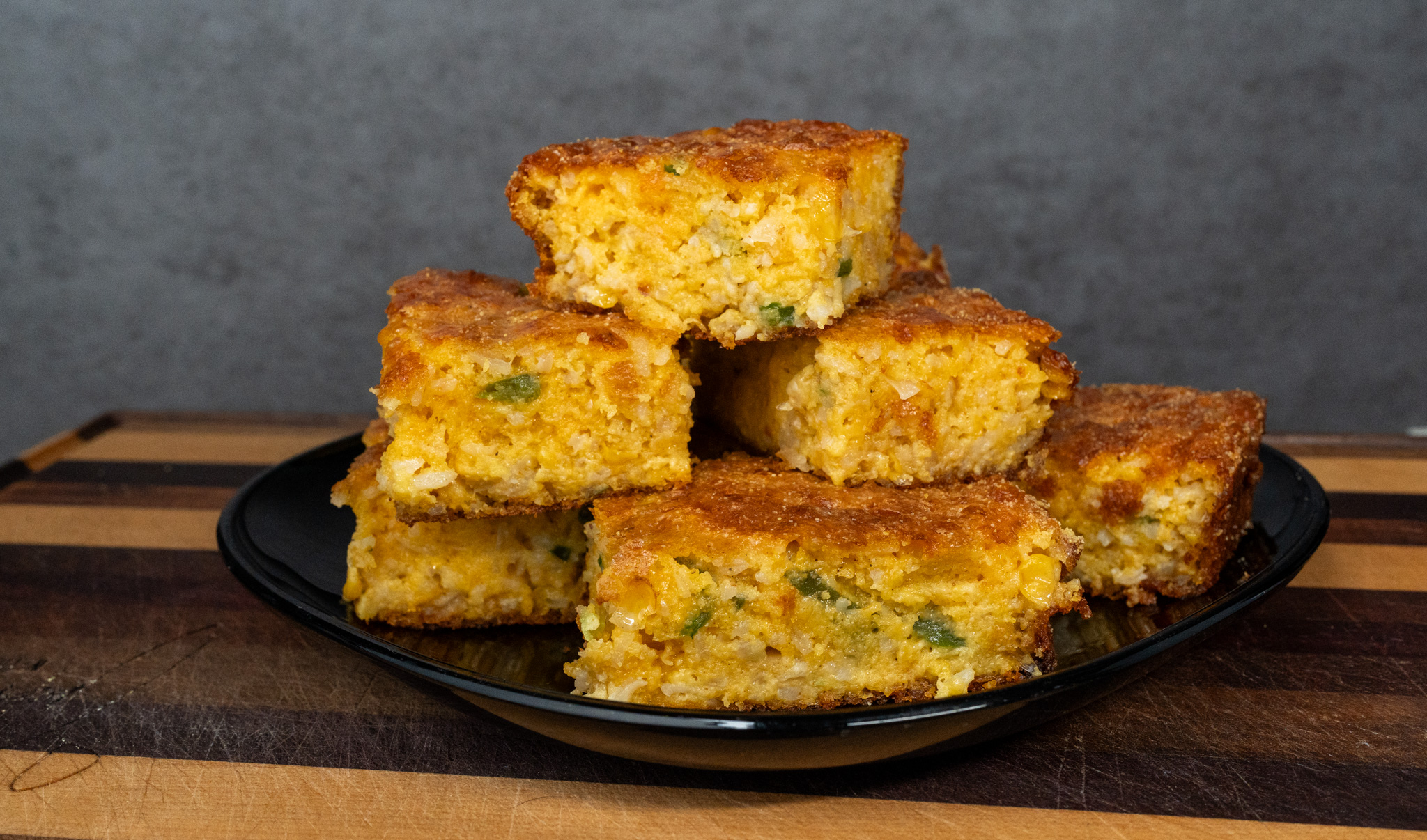 A stack of golden-brown Creole cornbread squares with visible pieces of green peppers, displayed on a black plate atop a wooden surface, set against a gray background.