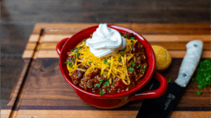 Bowl of homemade chili topped with shredded cheddar cheese, sour cream, and fresh parsley in a red dish on a wooden board.