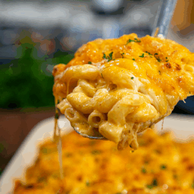A close-up of a spoon lifting a serving of cheesy southern mac and cheese from a casserole dish, with melted cheese stretching from the spoon.