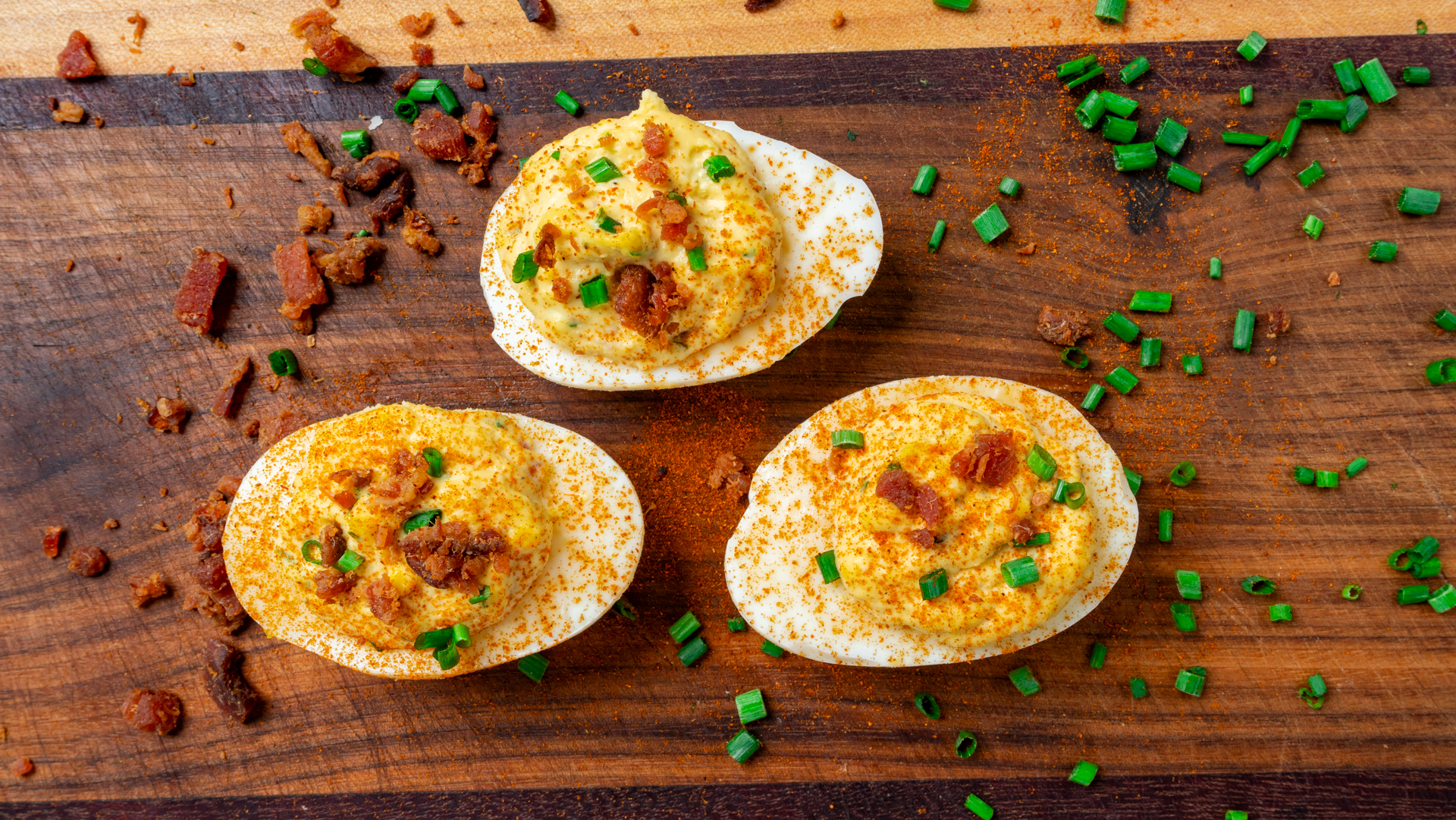 Three million dollar deviled eggs on a wooden board, topped with paprika, chopped chives, and crumbled bacon, with more chives and bacon sprinkled around them.