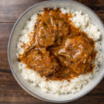 A bowl of white rice topped with three meatballs covered in a rich, brownish-red sauce—reminiscent of the flavors found in smothered oxtails—viewed from above on a wooden table.