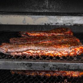 Two large pieces of brisket with a dark, crispy bark rest on a metal rack inside a smoker, smoked ribs could be prepared on the empty rack above them.