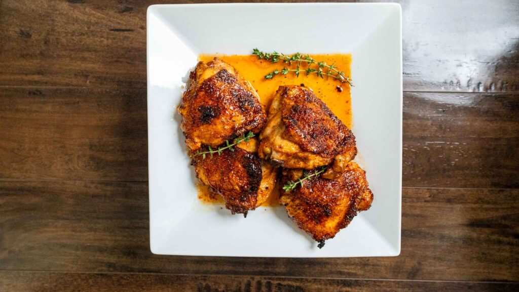 Four seasoned, baked hot honey chicken thighs with a golden-brown crust are arranged on a white square plate, garnished with fresh thyme sprigs and surrounded by a light orange sauce. The plate sits on a wooden table.