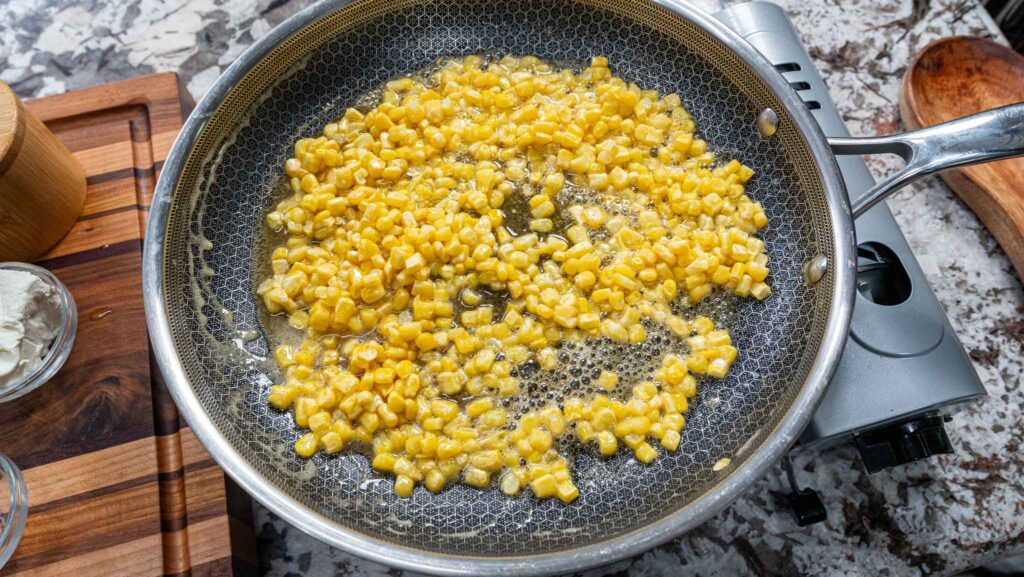 Fresh corn kernels sizzling in honey butter in a stainless steel skillet on the stovetop, with cooking ingredients and utensils arranged nearby on a wooden board.