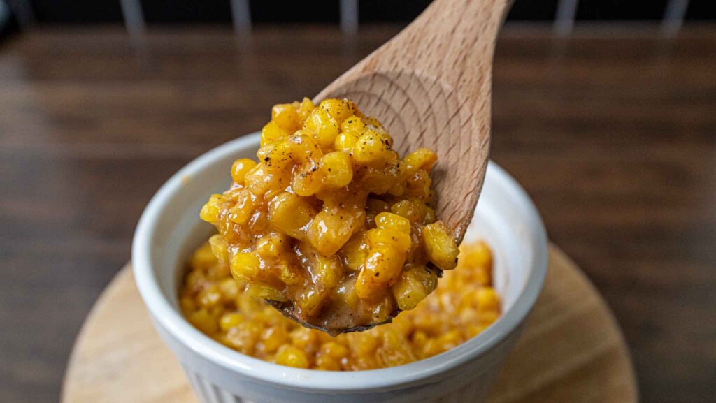 A wooden spoon holds a serving of creamy, seasoned honey butter skillet corn above a white ramekin filled with the same corn dish, set on a wooden surface.