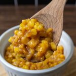 A wooden spoon holds a serving of creamy, seasoned honey butter skillet corn above a white ramekin filled with the same corn dish, set on a wooden surface.