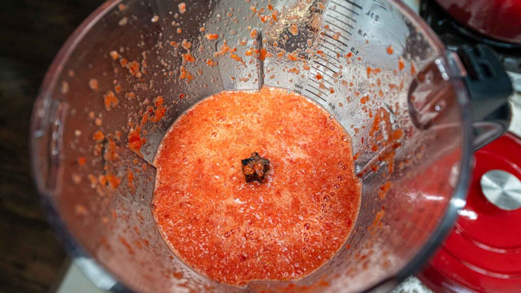 Blender container filled with a blended red mixture, likely used for making jollof rice or crushed fruit, with splatters on the sides and a visible black blade in the center.