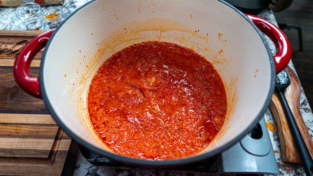 A large white pot with red handles contains simmering tomato sauce for jollof rice. The sauce is chunky and vibrant red, and the pot sits on a stovetop with a wooden cutting board and kitchen utensils nearby.