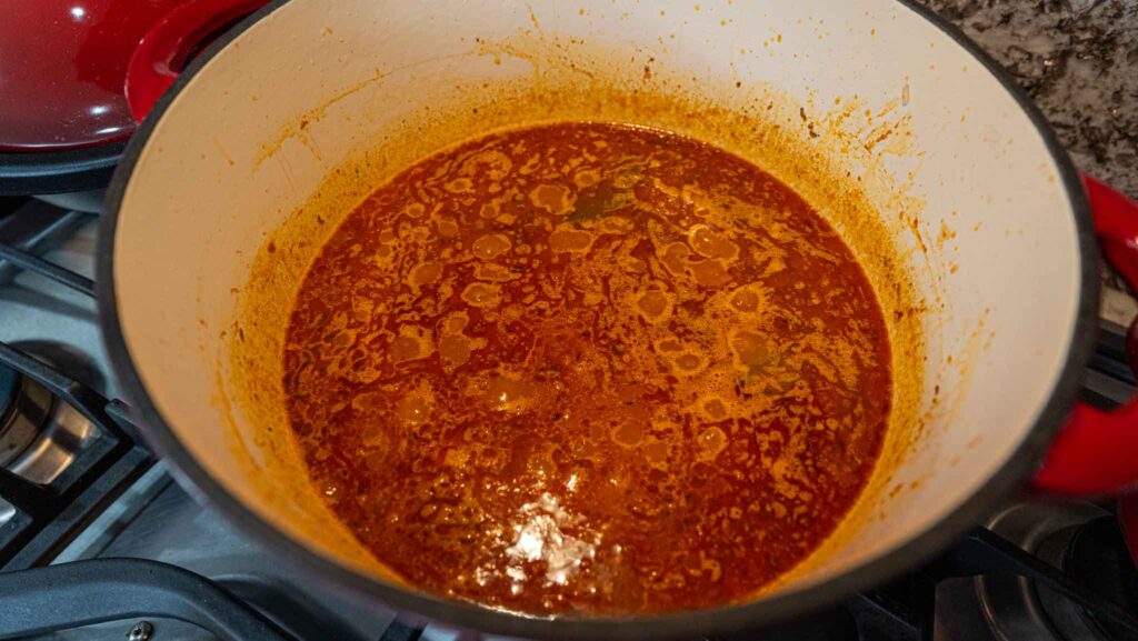 A red Dutch oven on a stove contains a simmering pot of reddish-brown jollof rice stew, with oil pooling on the surface and bits of food visible in the flavorful liquid.