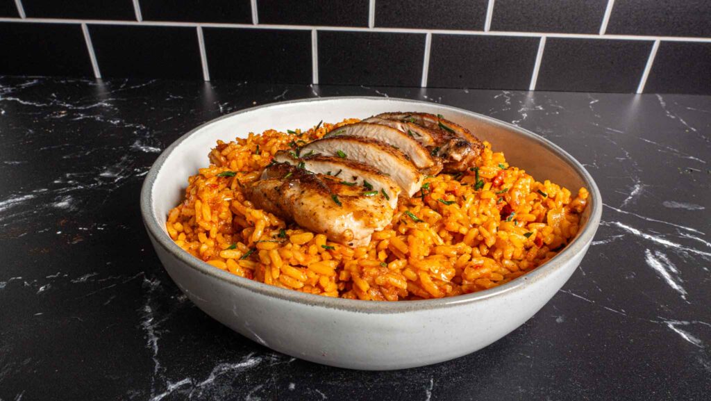A bowl of orange-colored jollof rice topped with sliced grilled chicken breast, garnished with herbs, sits on a black marble countertop with a black tiled wall in the background.