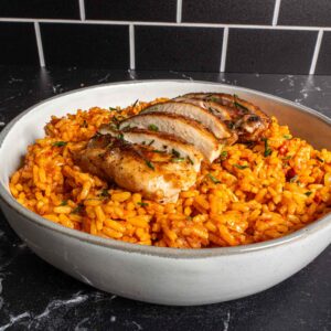 A bowl of orange-colored jollof rice topped with sliced grilled chicken breast, garnished with herbs, sits on a black marble countertop with a black tiled wall in the background.