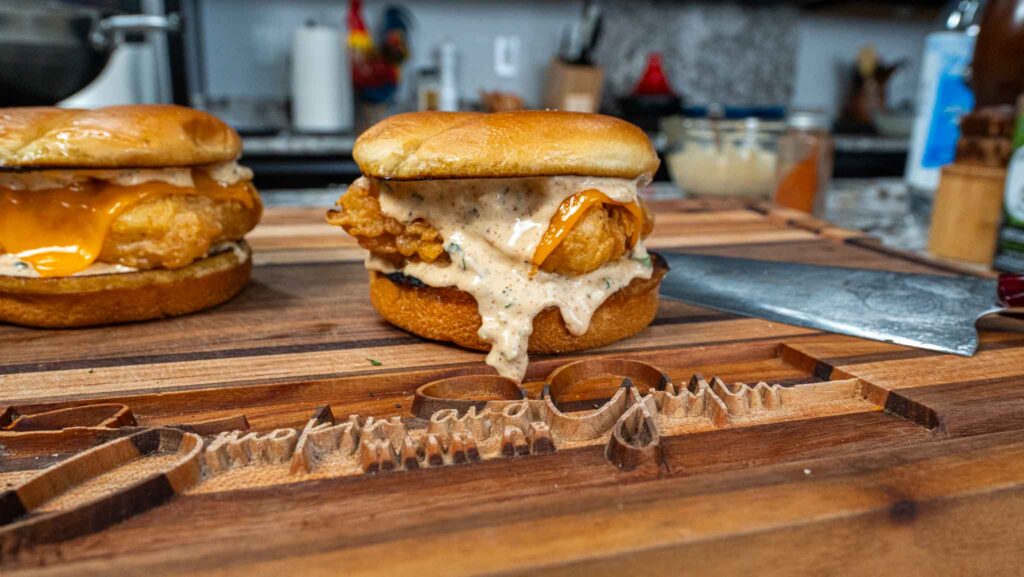 A fried chicken sandwich with melted cheese and creamy sauce sits on a wooden cutting board, next to another sandwich reminiscent of a filet o fish, and a knife, in a cozy kitchen setting.