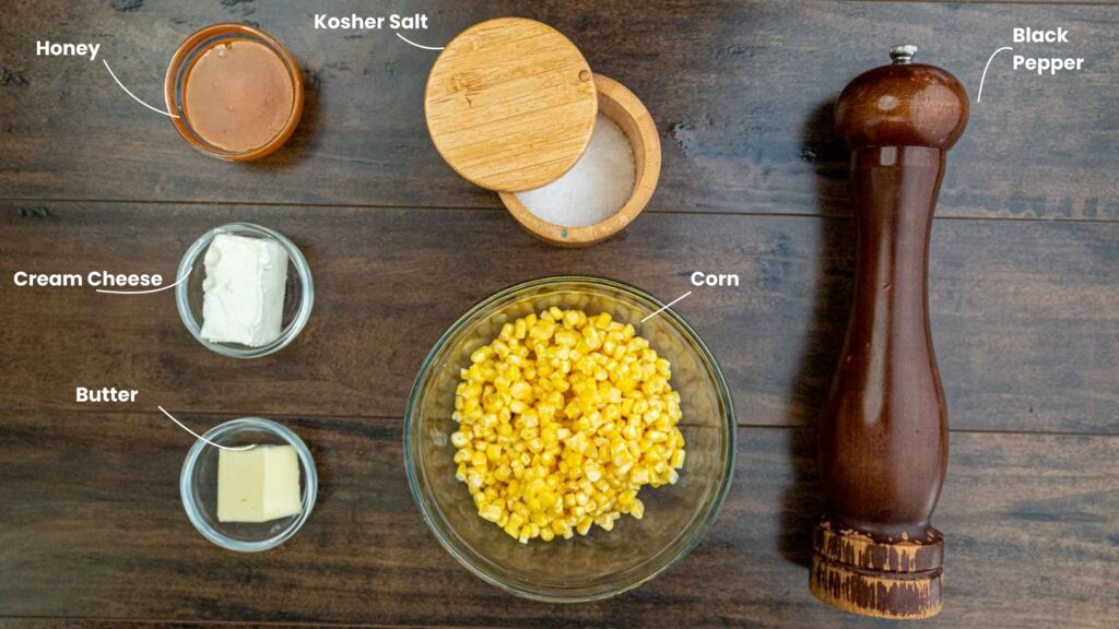 A top-down view of ingredients on a wooden table for honey butter skillet corn: a bowl of corn, cream cheese, butter, honey, kosher salt in a wooden container, and a large pepper grinder, each labeled accordingly.