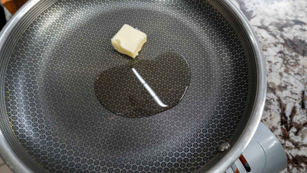A small cube of butter melting in a nonstick frying pan on a stovetop, ready to become the base for garlic butter asparagus, with some melted butter pooling beside it.