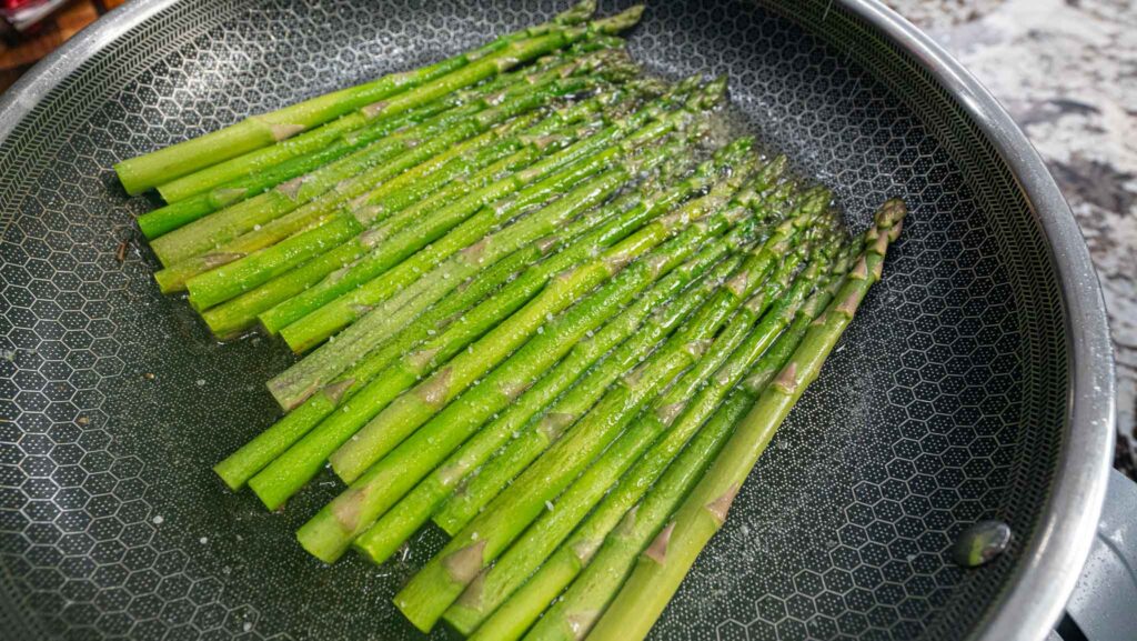 Fresh green asparagus spears cooking in a frying pan with garlic butter, lightly seasoned with salt and pepper.