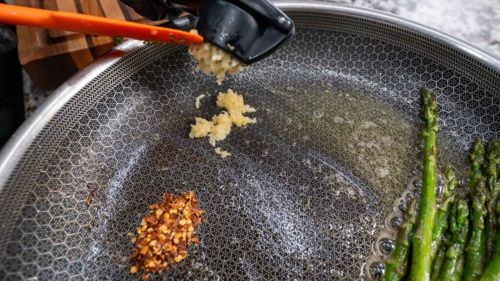 A garlic press adds minced garlic to a frying pan with hot oil, red pepper flakes, and tender spears of garlic butter asparagus.