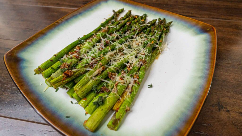 A plate of garlic butter asparagus spears garnished with grated parmesan cheese and herbs, arranged neatly on a decorative square plate with a green and brown rim, set on a wooden table.