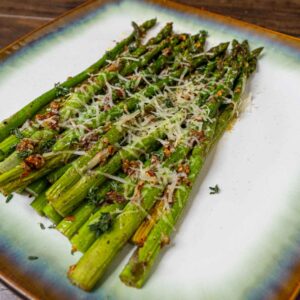 A plate of garlic butter asparagus spears garnished with grated parmesan cheese and herbs, arranged neatly on a decorative square plate with a green and brown rim, set on a wooden table.