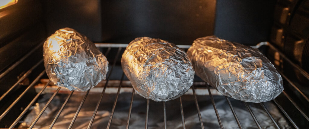 Three foil-wrapped Cajun baked potatoes are baking on an oven rack, with the oven door open and the spicy potatoes lined up side by side.