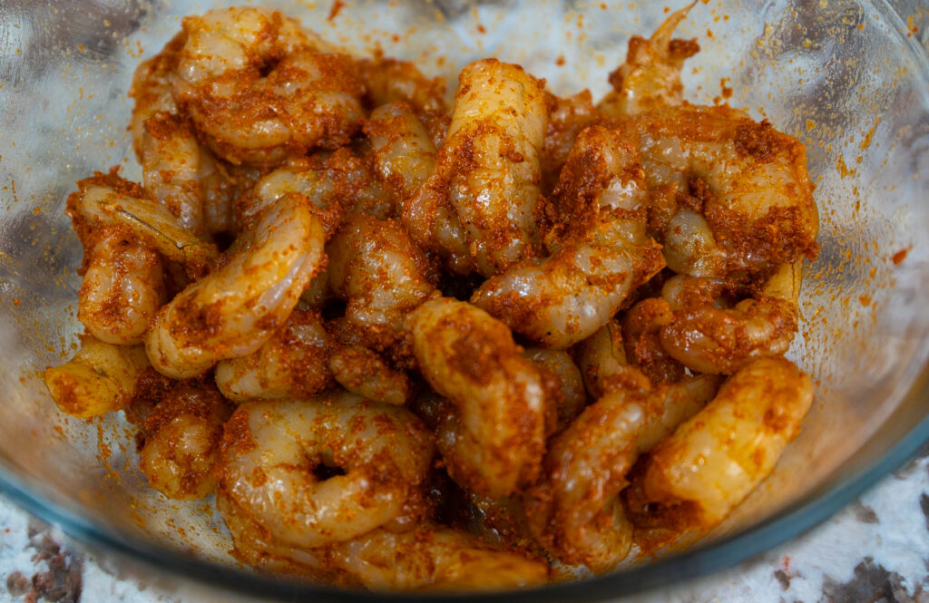 Raw shrimp coated with orange-red spices are in a glass bowl, ready for cooking. The shrimp are deveined and appear marinated, similar to the bold seasoning found on a cajun baked potato, with visible spices clinging to their surfaces.