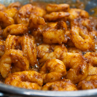 Shrimp cooking in a metal skillet, coated with a rich, orange-brown sauce and seasonings, alongside cajun baked potato for a flavorful pairing. The shrimp are partially caramelized for added depth.