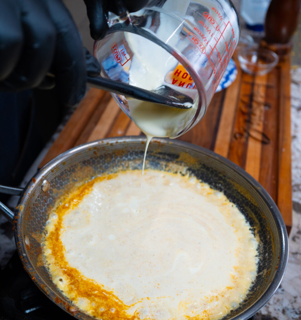 A person wearing black gloves pours a creamy liquid from a measuring cup into a pan with yellow-orange cajun baked potato sauce on a wooden countertop.