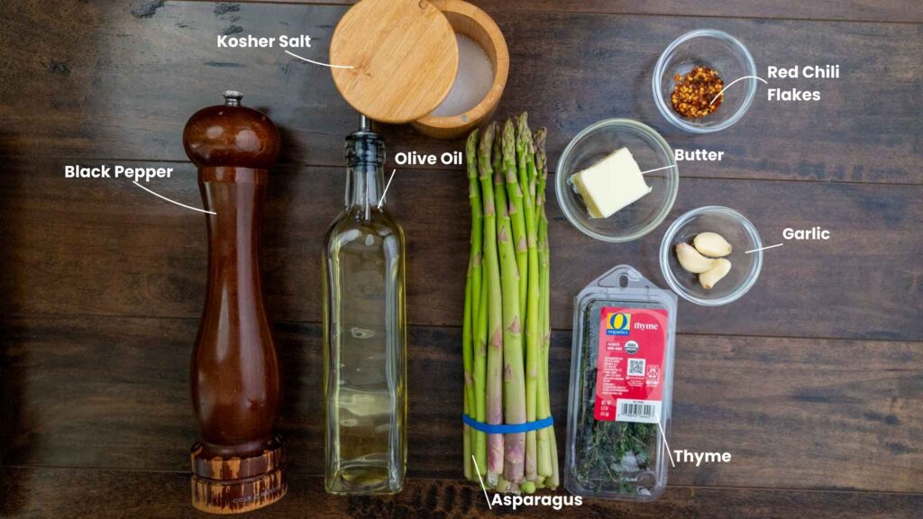 A flat lay of garlic butter asparagus ingredients—olive oil, black pepper grinder, kosher salt, butter, garlic cloves, red chili flakes, and a thyme packet—all labeled on a dark wooden surface.