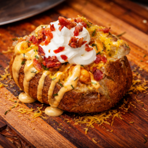 A Cajun baked potato topped with melted cheese, chopped bacon, sour cream, and creamy sauce, served on a wooden board with shredded cheese scattered around.