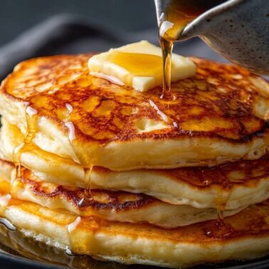 A stack of fluffy pancakes, just like granny's Homemade Hotcakes, is topped with a pat of butter and drizzled with syrup from a small pitcher, served on a black plate against a dark background.