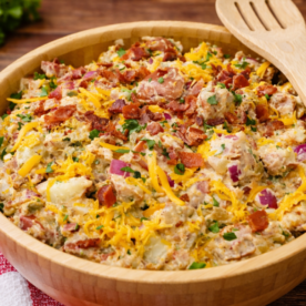 A wooden bowl filled with Steakhouse Potato Salad—loaded with bacon bits, shredded cheese, and chopped chives—sits on a wooden table beside a spoon, salt and pepper shakers, and a red checkered napkin. Perfect for learning how to make steak house potato salad!.