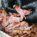 A person wearing black gloves is pulling apart smoked, shredded meat over a tray of tender, juicy barbecue—showcasing the results of mastering how to smoke pork butt with pecan pellets.