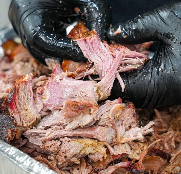 A person wearing black gloves is pulling apart smoked, shredded meat over a tray of tender, juicy barbecue&mdash;showcasing the results of mastering how to smoke pork butt with pecan pellets.