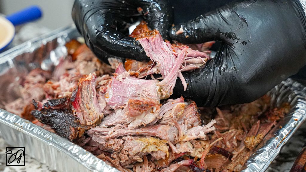 A person wearing black gloves is pulling apart smoked, shredded meat over a tray of tender, juicy barbecue—showcasing the results of mastering how to smoke pork butt with pecan pellets.