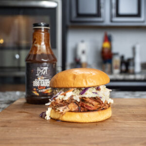 A pulled pork sandwich with coleslaw on a wooden board, featuring tender meat prepared using the How to Smoke Pork Butt with Pecan pellets method. A bottle of BBQ sauce and a kitchen with dark cabinets complete the scene.